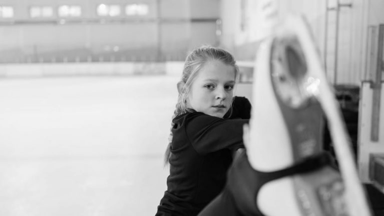 A focused girl practicing figure skating on an indoor ice rink in monochrome.